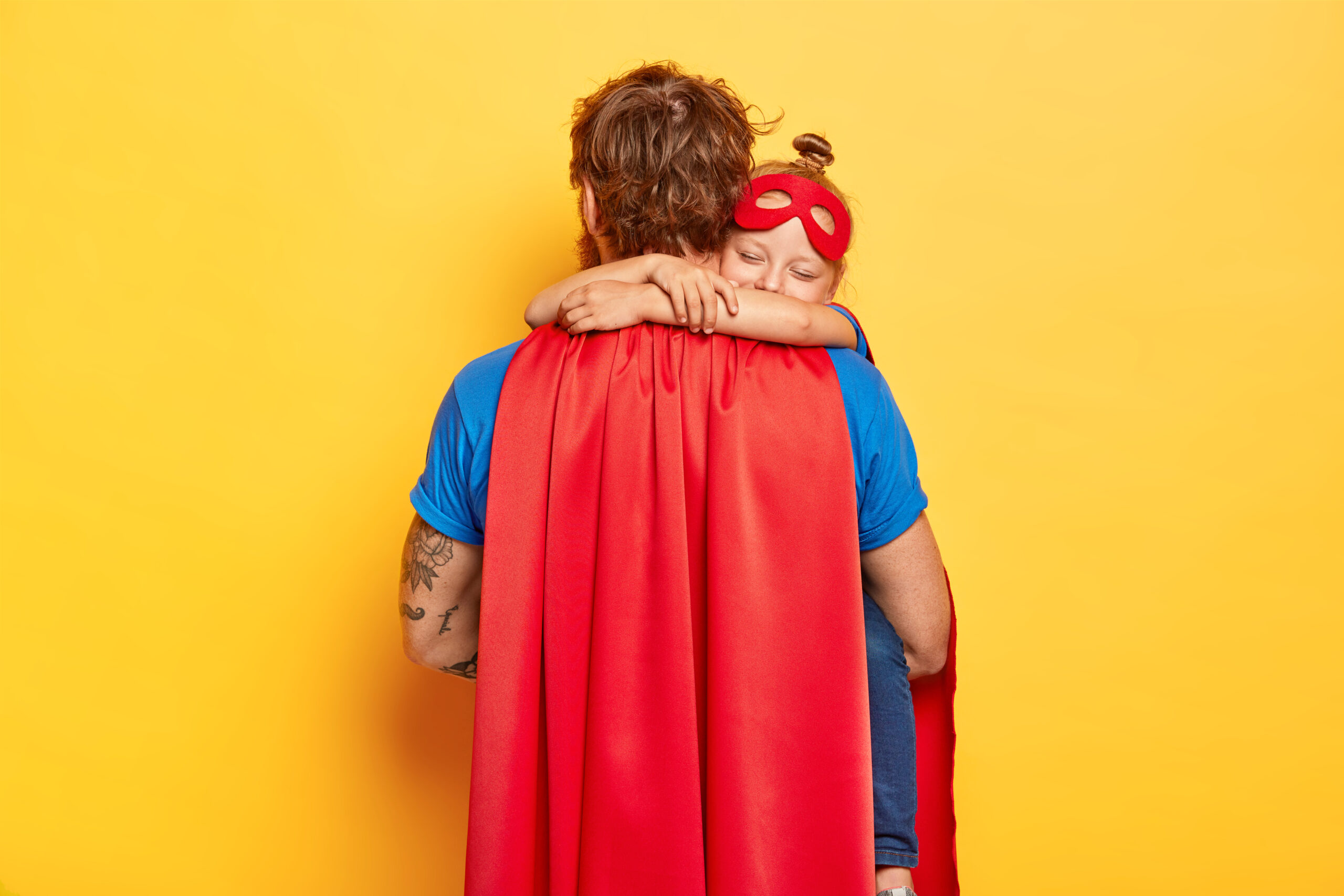 Horizontal shot of caring father carries small daughter, receives warm hug, wear red cloak and mask, play superhero game, dressed in special costumes, isolated on yellow wall. Male defender with kid